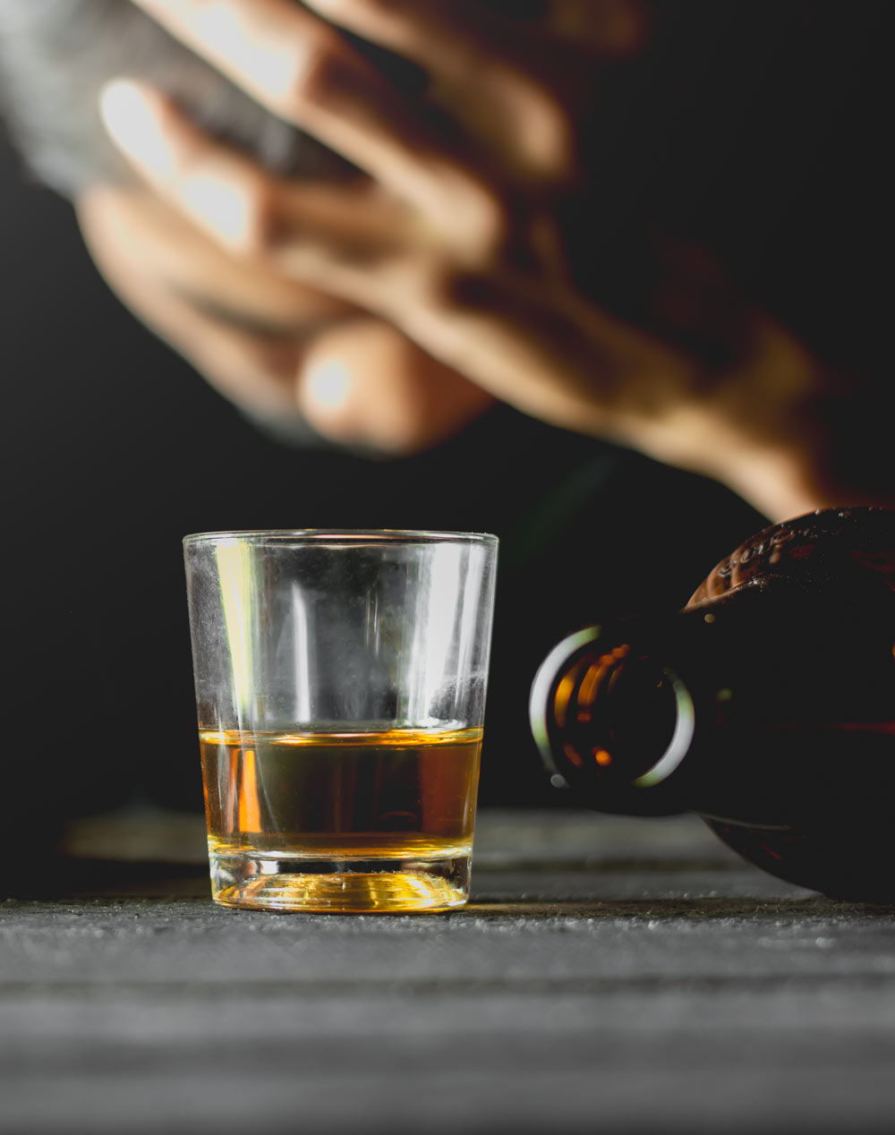 man holding his head leaning down with a shot of alcohol in front of him