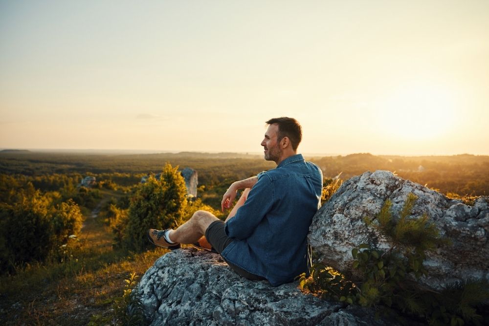 A man sits on a large rock at sunset, gazing peacefully over a vast green landscape. He looks thoughtful and relaxed, enjoying the quiet view.