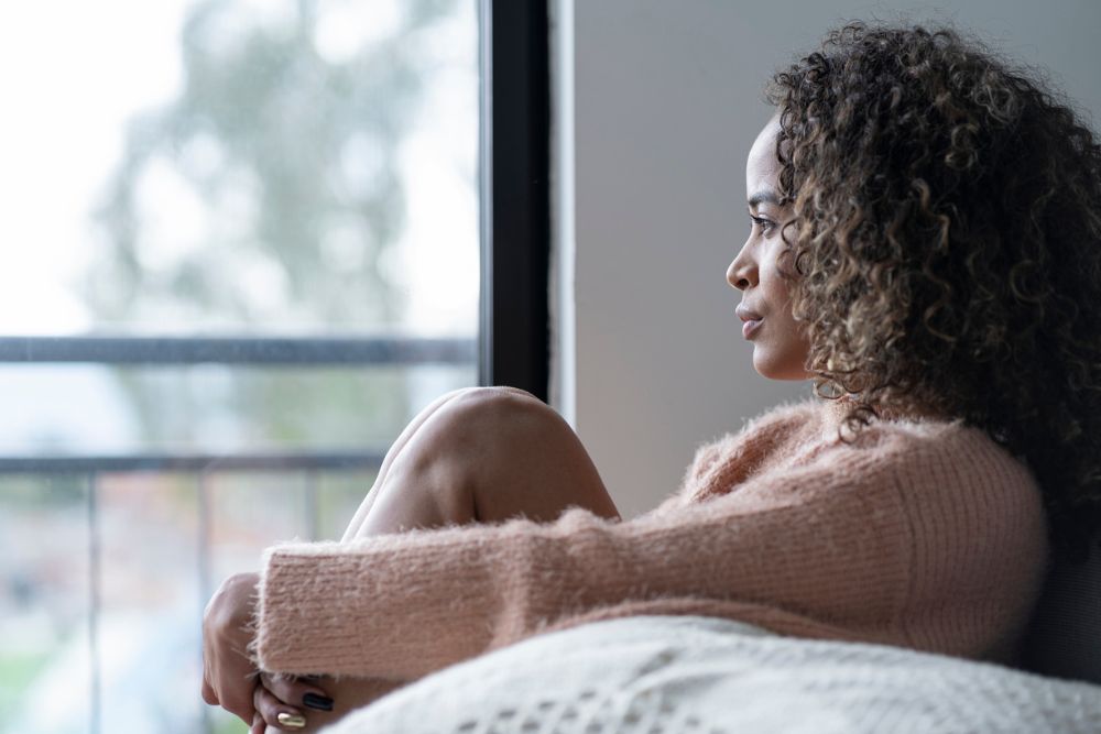 A woman sits curled up on a couch, looking out a window with a serious expression, suggesting deep thought, emotional pain, or reflection.