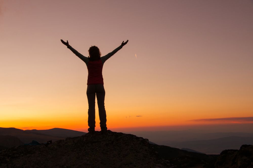 Silhouette of a person standing on a mountaintop at sunrise with arms raised, embracing the colorful sky and expansive horizon.