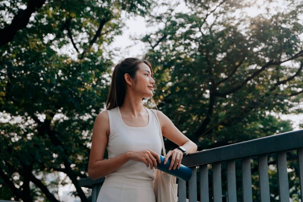 Woman in athletic wear holding a water bottle, leaning on a railing and peacefully looking into the distance, surrounded by trees and natural light.