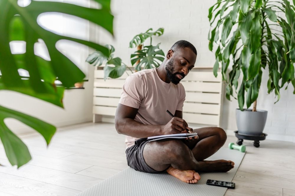 A man sits cross-legged on a yoga mat writing in a notebook, surrounded by plants and light, symbolizing mindfulness and healthy habits.