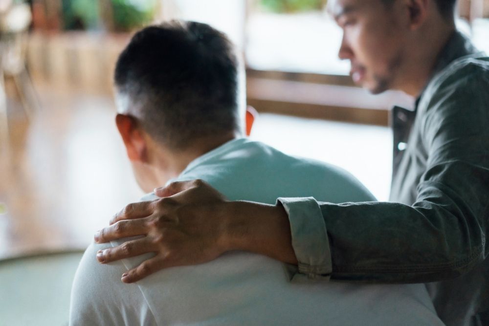 A man rests his hand on another man’s shoulder in a gesture of comfort and support.