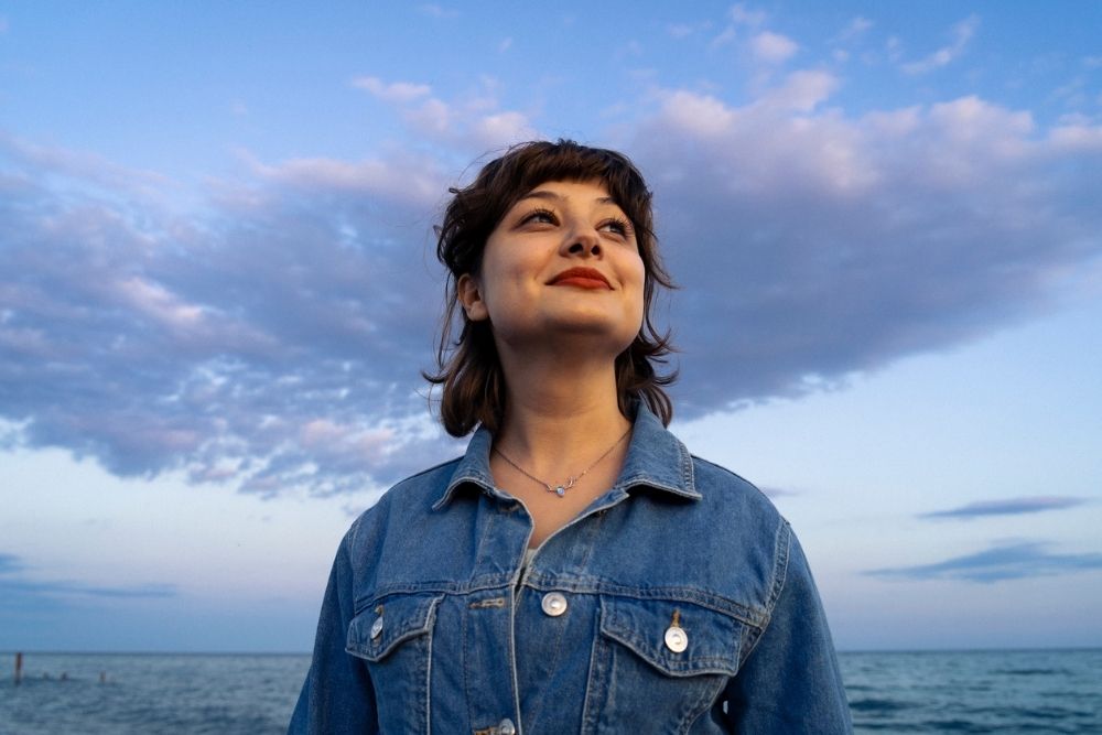 Woman standing outdoors by the water, looking hopeful and smiling slightly under a blue sky with clouds.