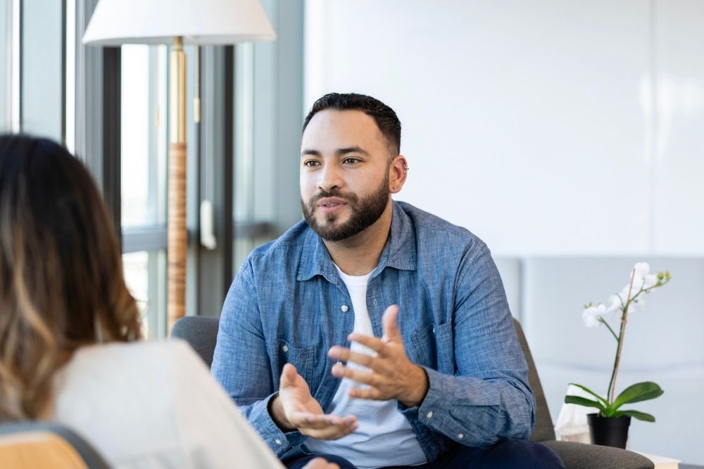 A man speaks with a therapist during a counseling session, gesturing as he talks in a calm, professional office.
