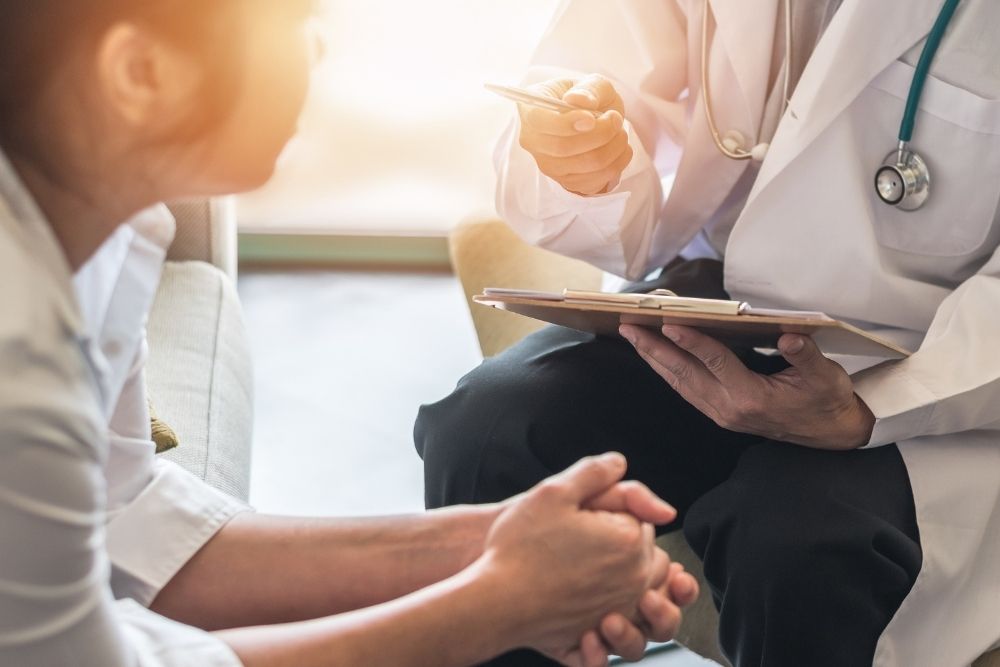 A healthcare professional holding a clipboard and speaking with a seated patient during a medical consultation.