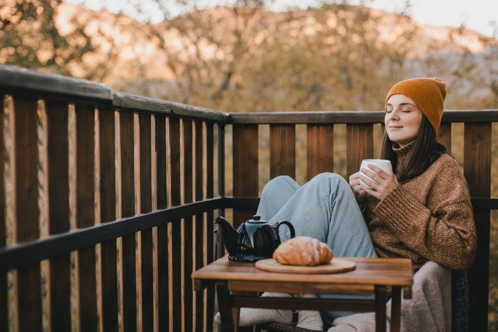 A woman sitting on a cozy outdoor deck in fall, holding a warm drink with her eyes closed, enjoying a peaceful moment beside a teapot and pastry.