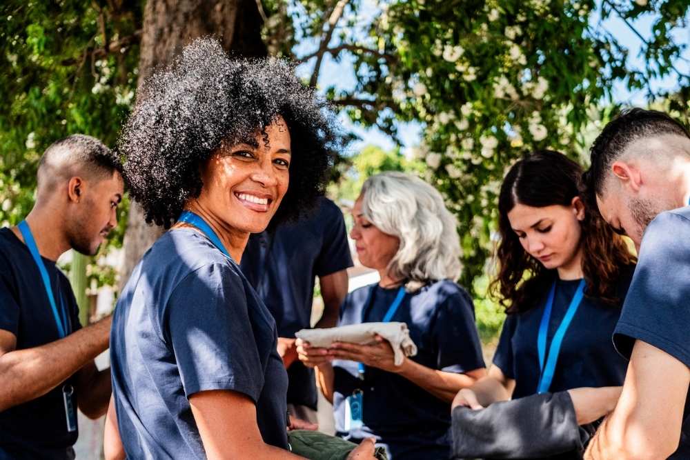 A diverse group of people wearing navy shirts and lanyards engage in outdoor volunteering work. A woman in the foreground smiles warmly, creating a positive and collaborative atmosphere.