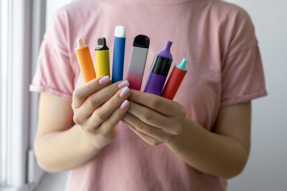 A person in a pink shirt holds seven colorful vapes in both hands, showcasing an array of brightly-colored e-cigarettes.